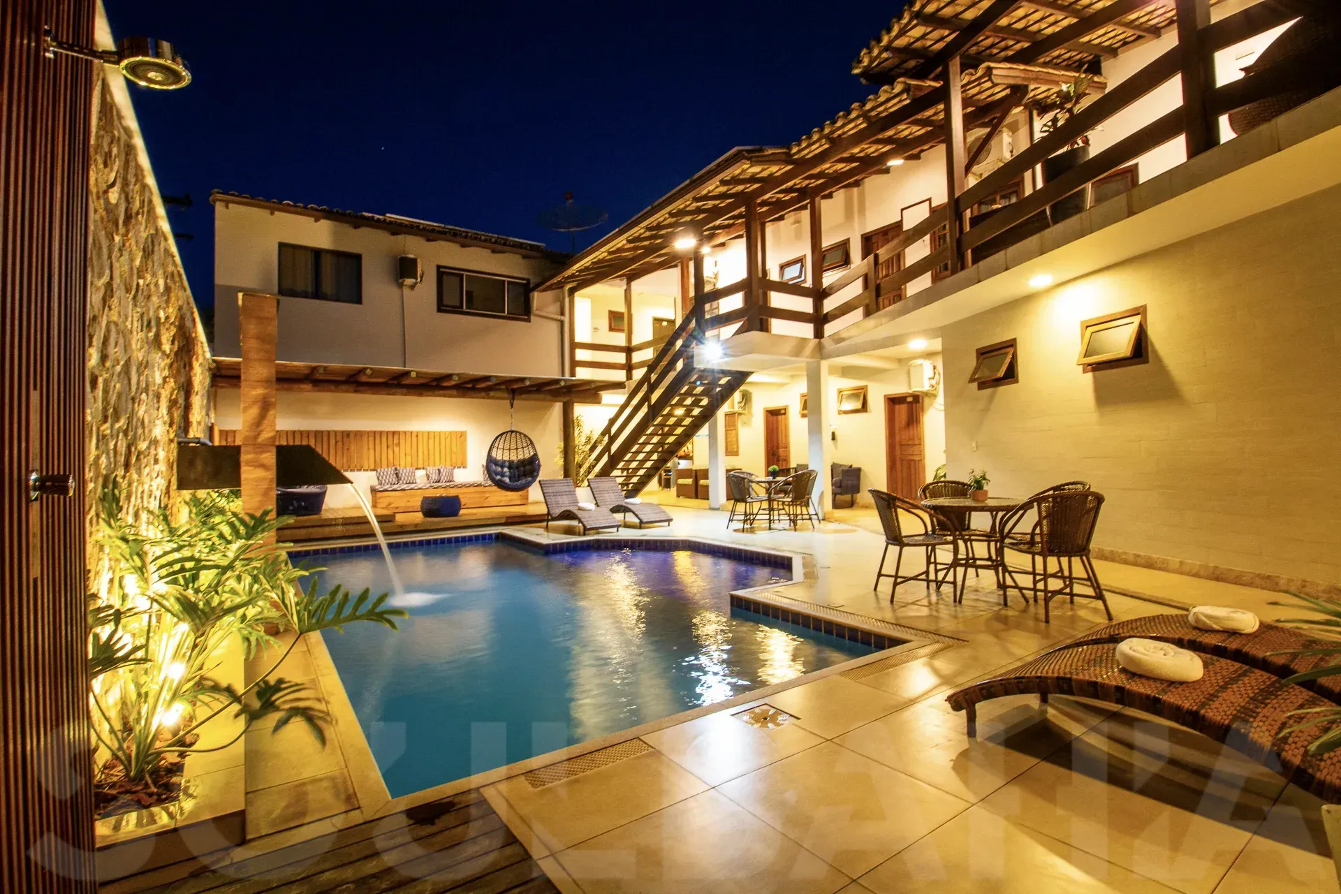 Piscina iluminada no pátio com espreguiçadeiras e varanda de dois andares à noite.