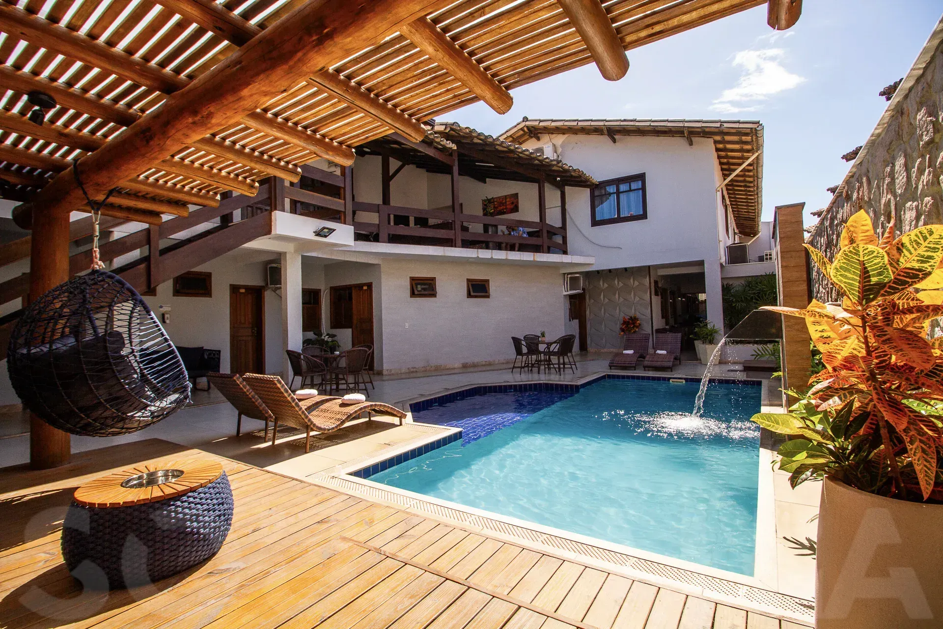 Pátio coberto com vista para uma piscina azul ao lado de uma casa branca com espreguiçadeiras de madeira e decoração em vime.