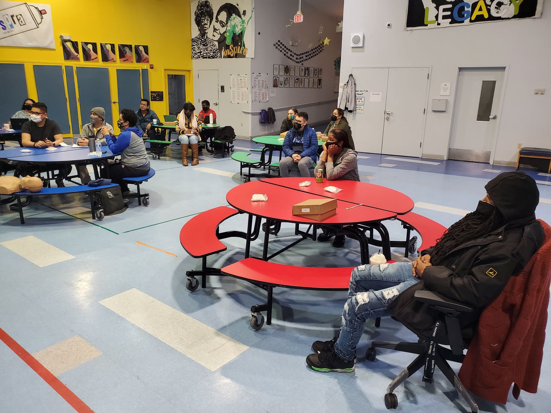 A group of people are sitting at picnic tables in a gym.