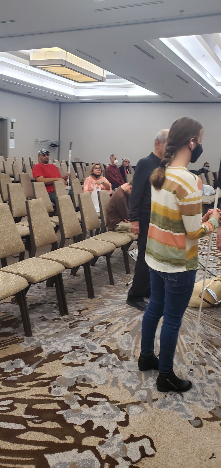 A woman wearing a mask is standing in a room with people sitting in chairs.