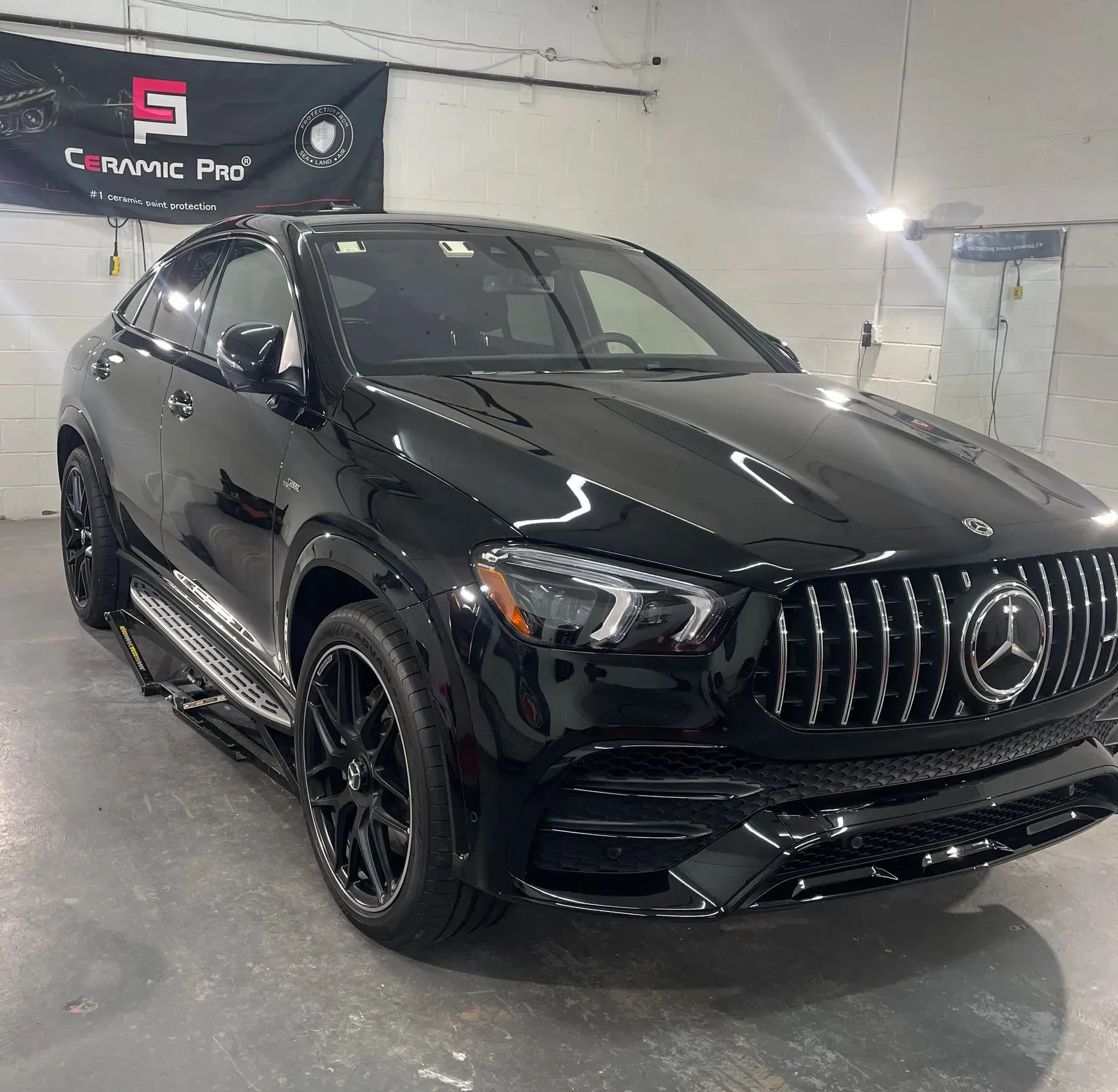 A shiny black Mercedes-Benz SUV parked inside a garage with a Ceramic Pro banner visible in the background.