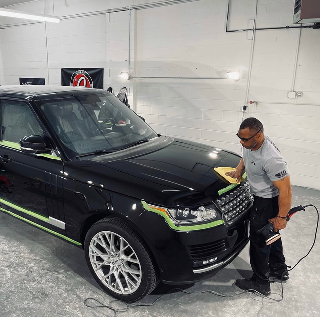A person in an auto shop uses a buffer to polish the hood of a black SUV with green detailing tape applied to its body.