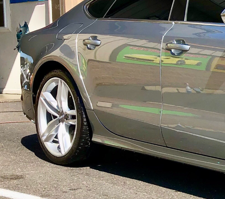 A close-up side view of a gray car featuring a silver alloy wheel and reflections of a yellow vehicle on the door.