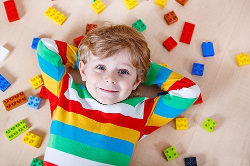 A young boy is earing a colorful shirt and laying on the ground with colorful Legos surrounding him.