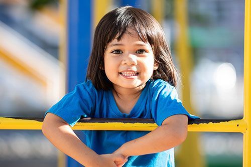 Young child with dark hair smiles at the camera, leaning on yellow playground equipment, wearing a blue shirt.