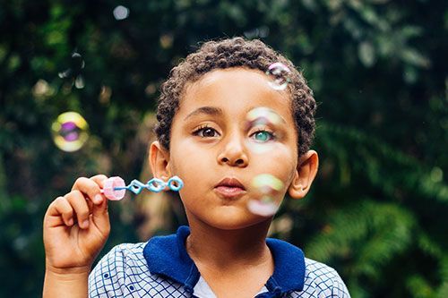 Boy blowing bubbles; pink wand, blue shirt, green background.