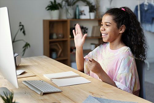 Girl waving at computer screen, smiling, at a wooden desk with a notebook and keyboard.