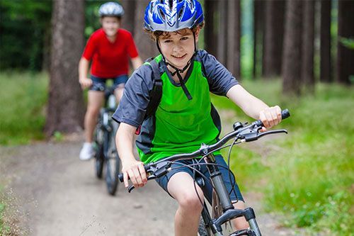 Two children riding bikes on a wooded path, wearing helmets. Boy in green shirt smiles.