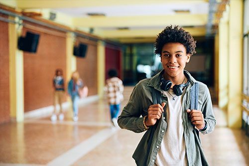 Teenage student in a school hallway smiles, wearing backpack and headphones.