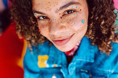 Person with curly hair, freckles, and colorful face gems smiles in a blue denim jacket.