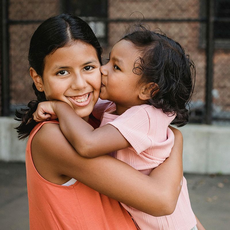Two children embracing outdoors; one kisses the other on the cheek, both smiling.
