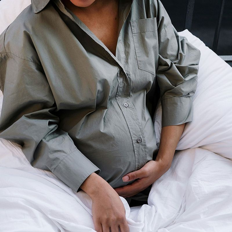 Pregnant person in gray button-down shirt resting in white bed, hands on belly.