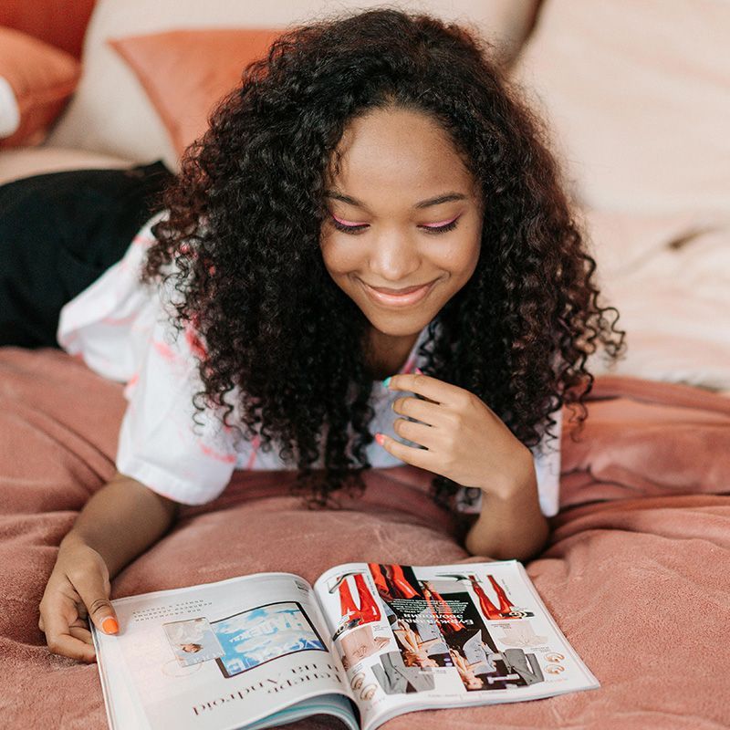 Woman with curly hair smiles while reading a magazine on a bed with pink linens.