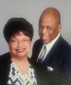 Two people, a woman and a man, smiling and posed formally. The man wears a suit with a striped tie. The woman wears glasses and a patterned blouse.