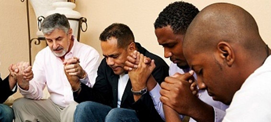 Group of men with clasped hands, heads bowed in prayer. Soft lighting, neutral background.