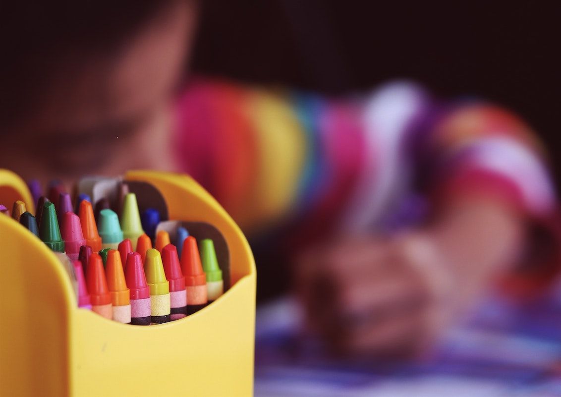 Yellow crayon holder with colorful crayons, child in rainbow shirt in background.