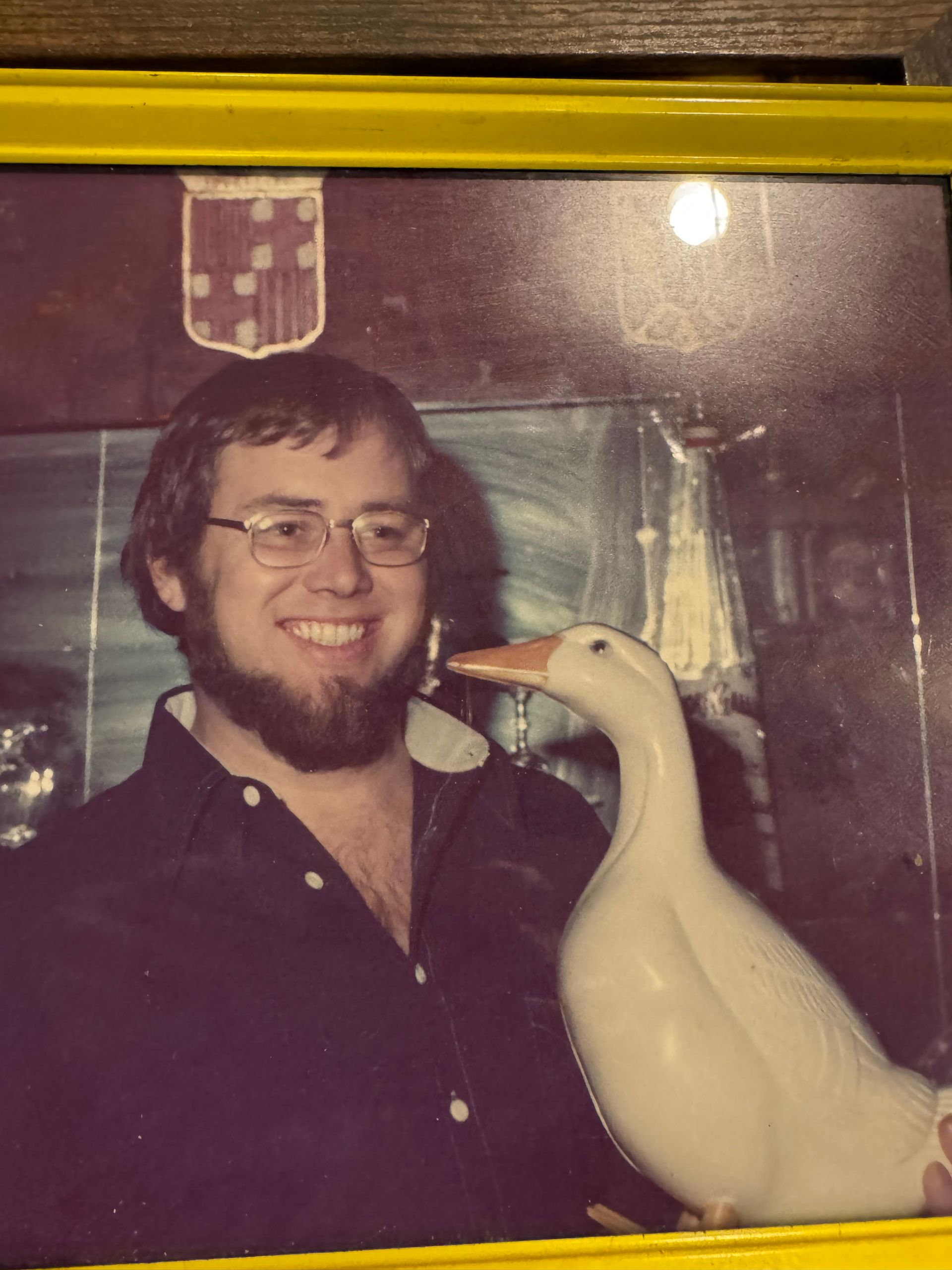A smiling person with a beard and glasses poses beside a large white decorative goose statue.