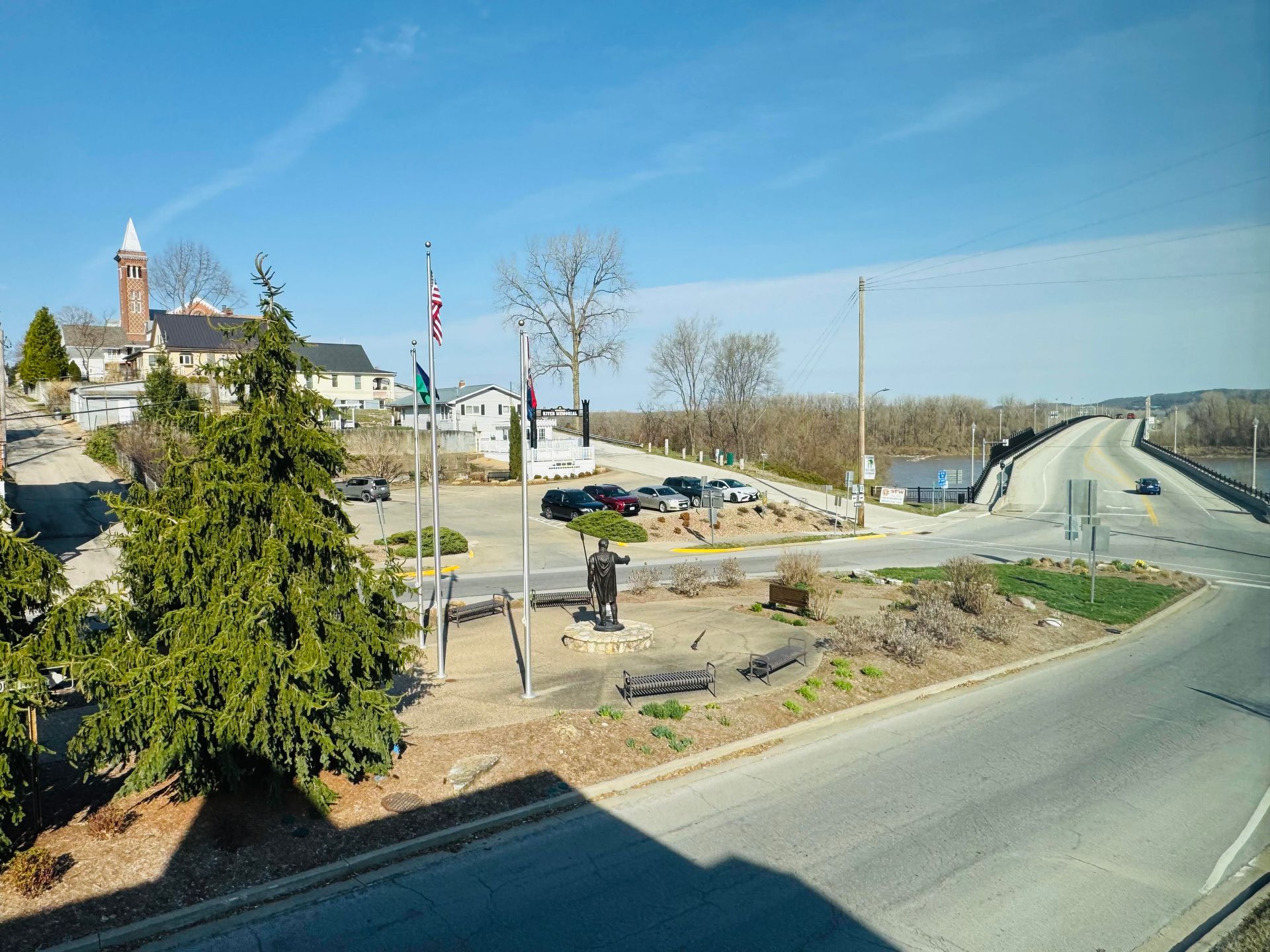 A sunny day at a small park with a statue, flagpole, and a bridge leading over water toward a hilltop building.