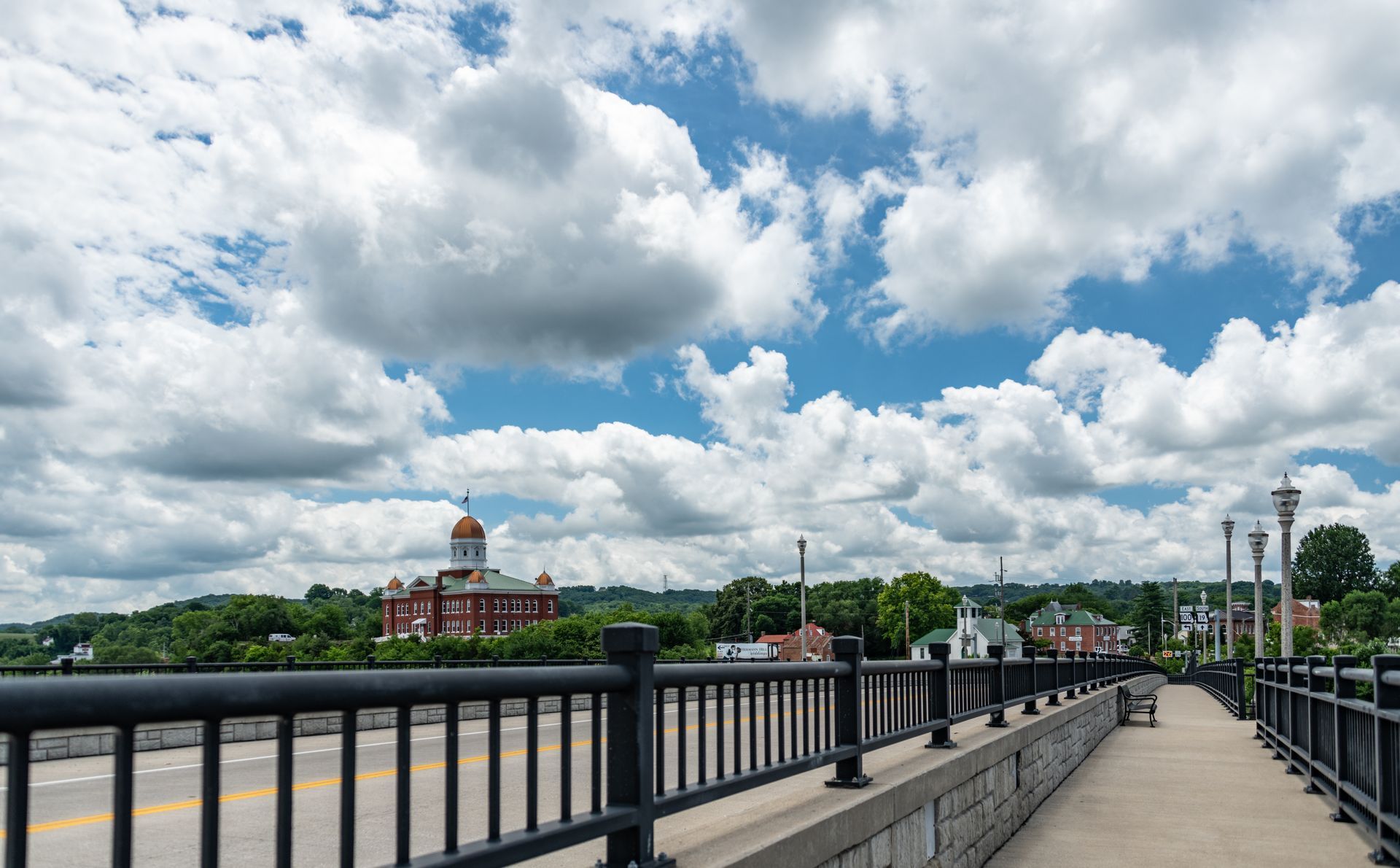 A perspective view of a bridge sidewalk with black railings, overlooking a small town under a cloudy blue sky.