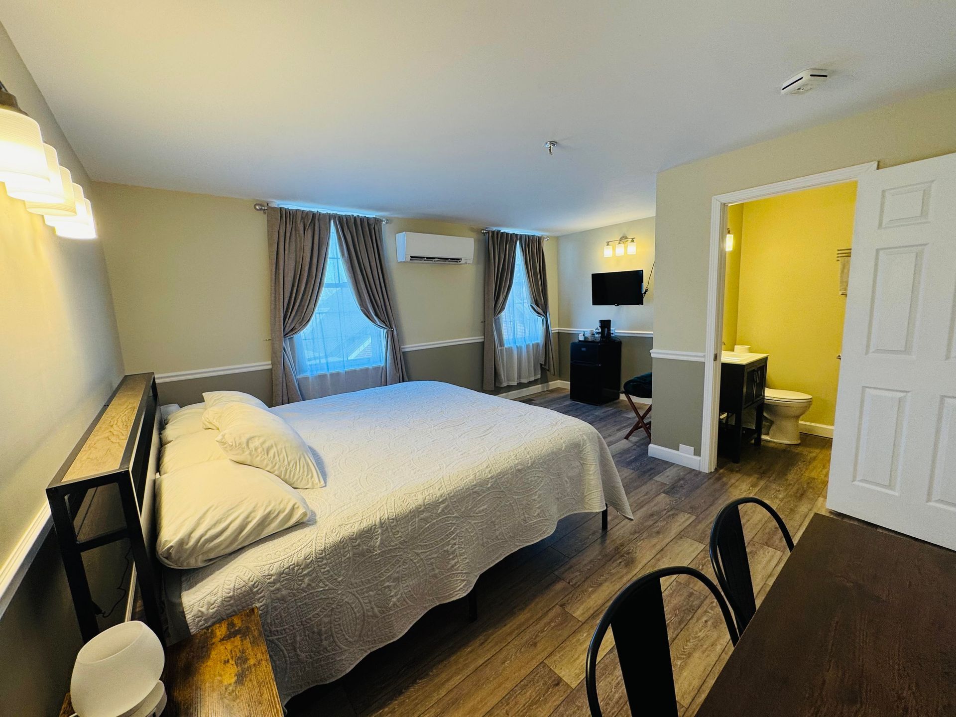 A hotel room with a queen bed, two windows with brown curtains, a small desk, a television, and an open bathroom doorway.