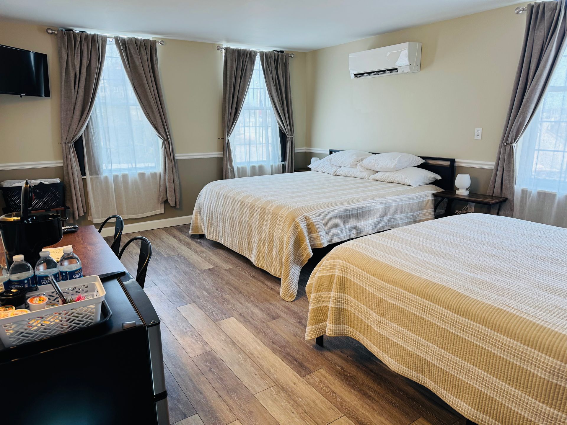 A hotel room with two queen beds, wood-look flooring, light walls, and a small table with supplies in the foreground.