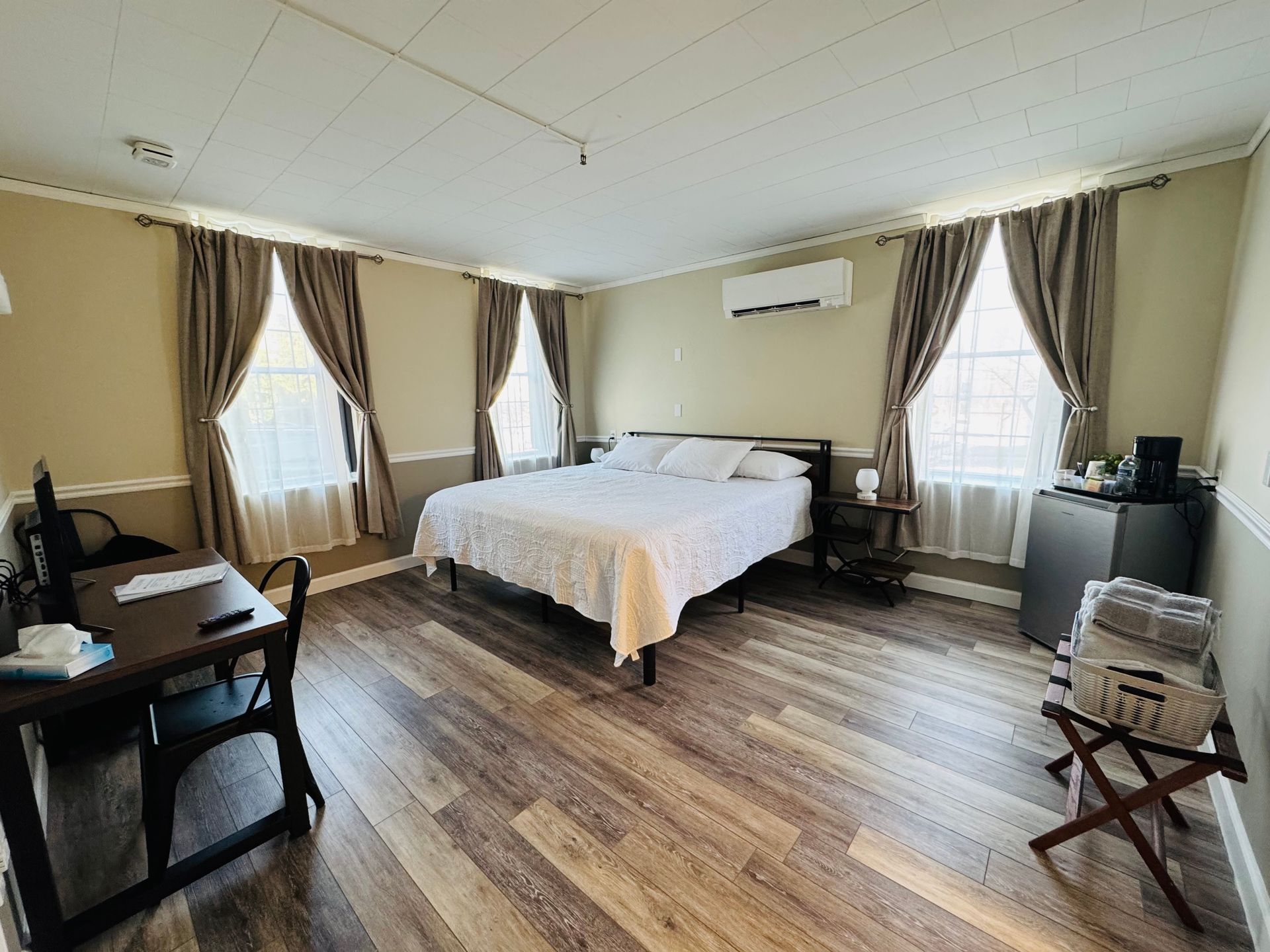 A bright, neutral-toned hotel room featuring a queen bed, wooden floors, two windows with curtains, and a desk.