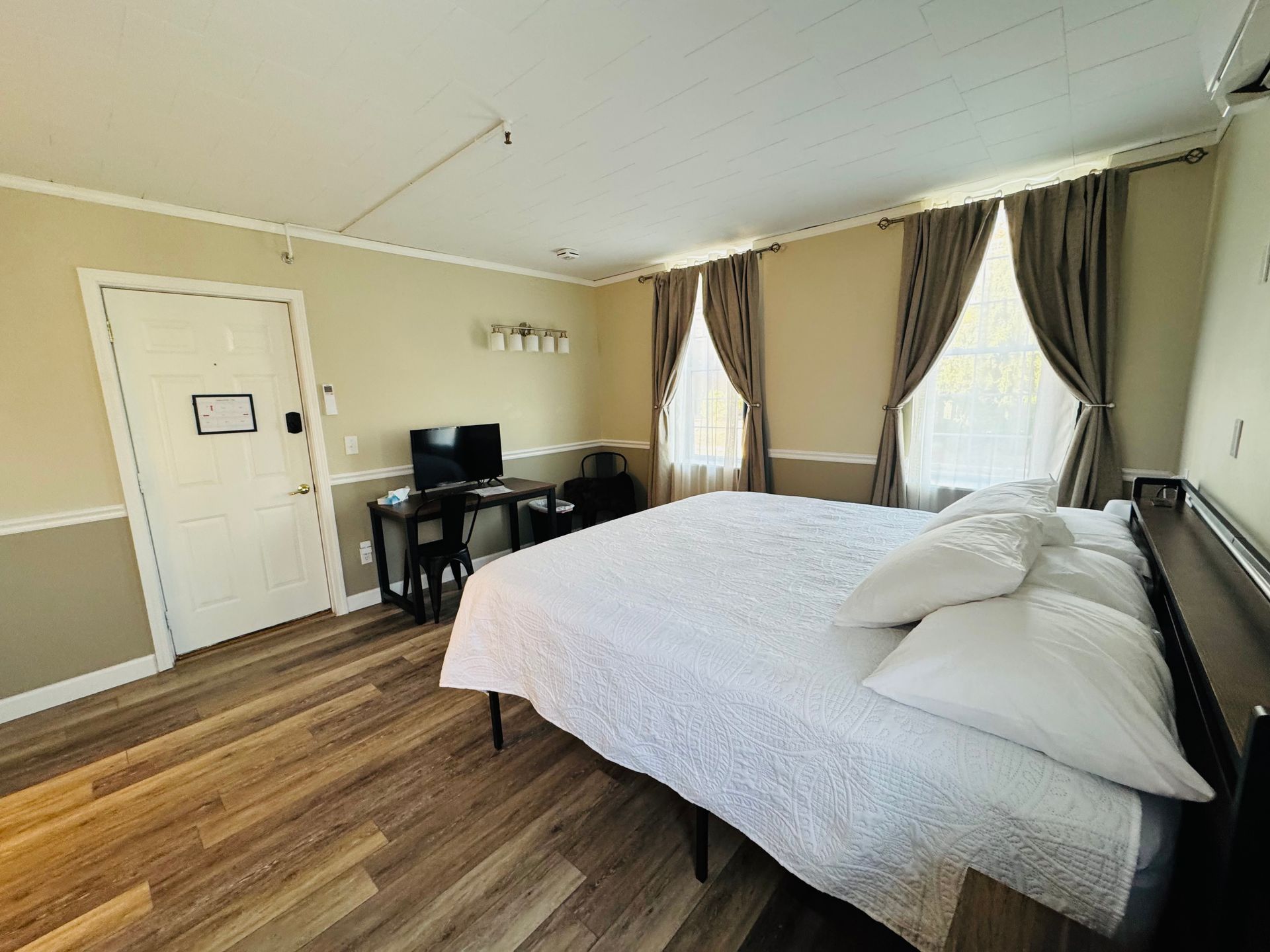 A hotel room with a large bed, light wood floors, a desk with a television, and floor-length curtains over two windows.