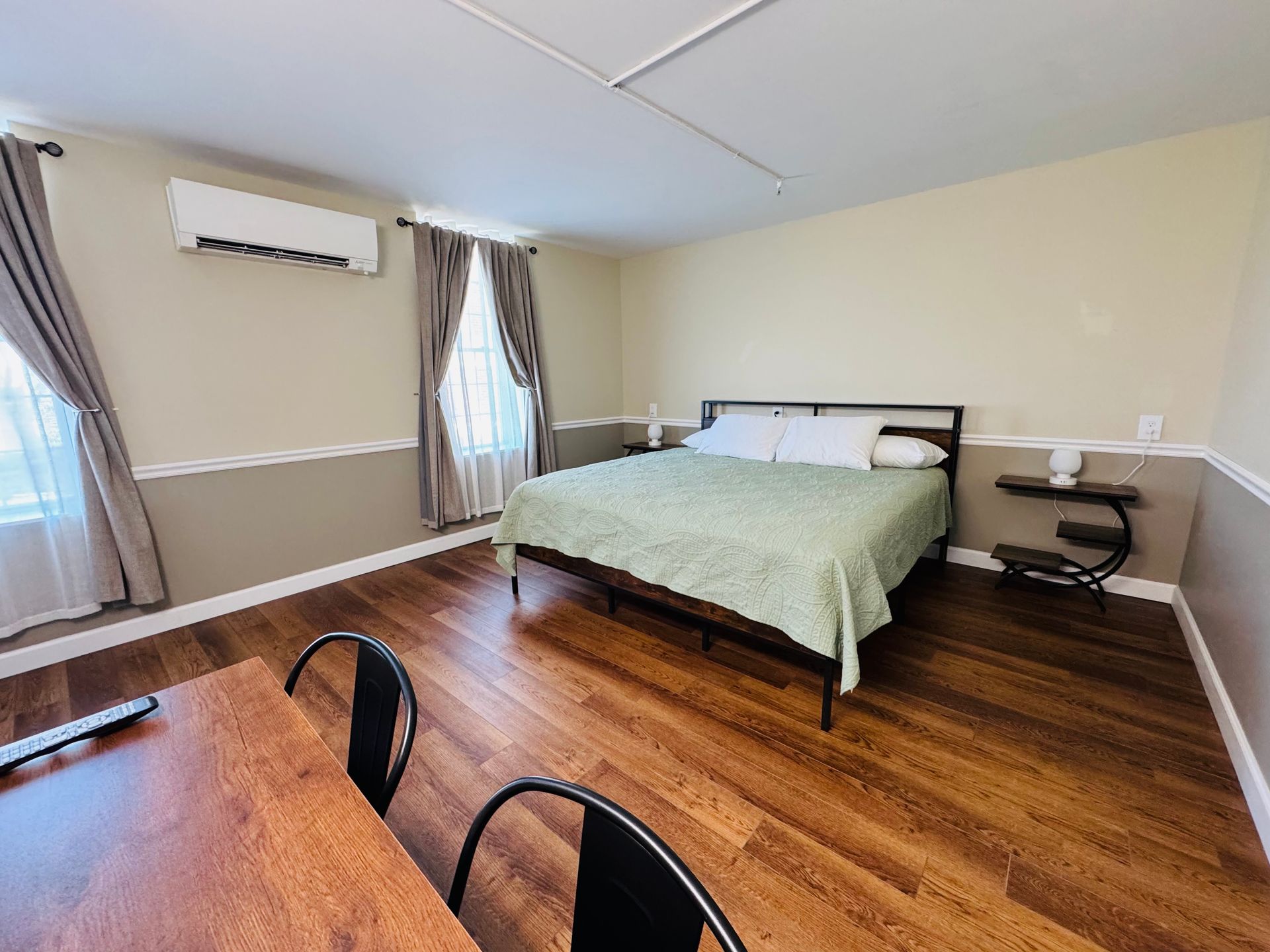 A bedroom with a king bed, light green quilt, wood floors, two windows with curtains, and a side table on the right.