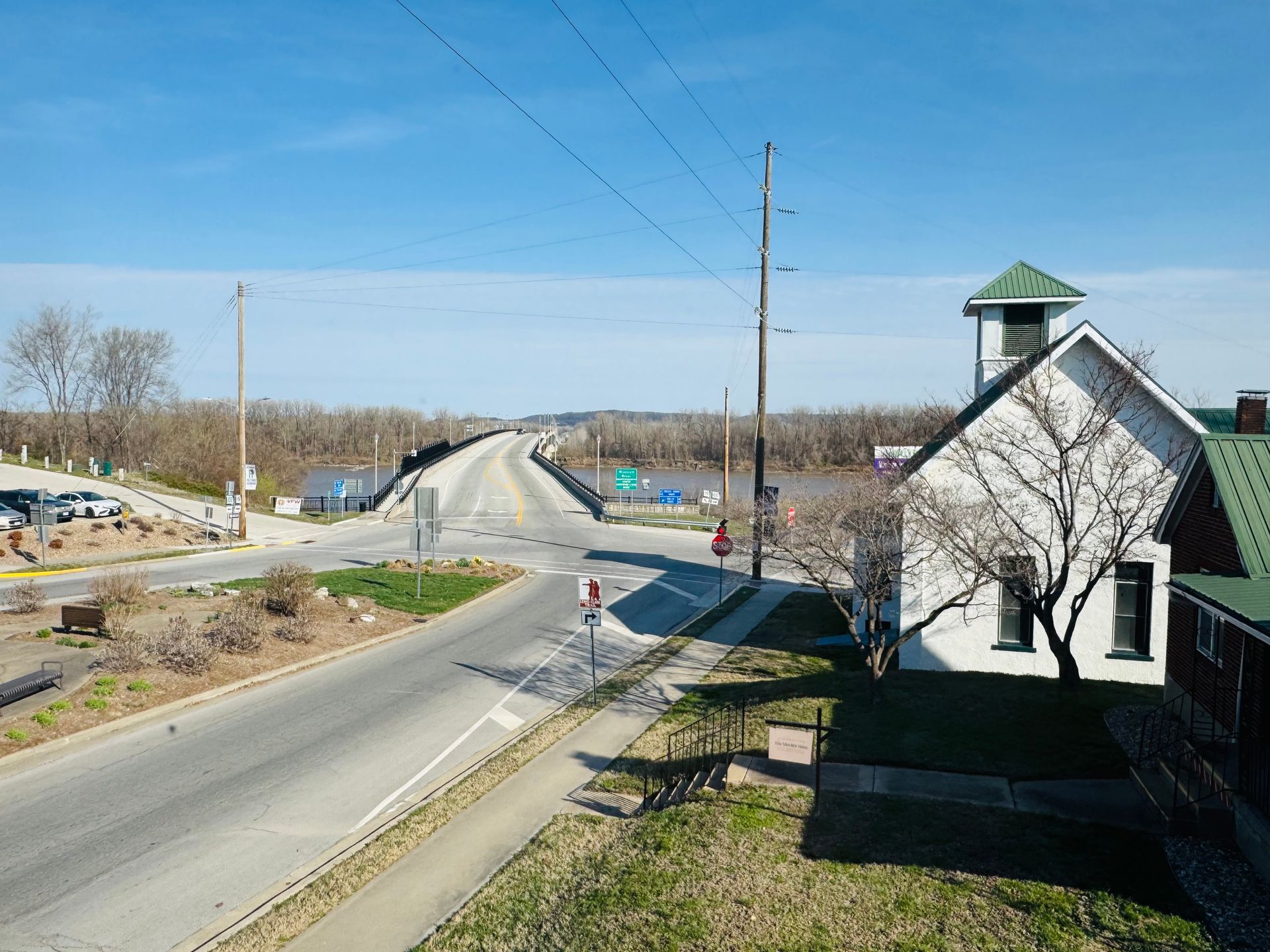 A bridge spans a wide river near a white building with a green roof on a sunny day under a clear blue sky.