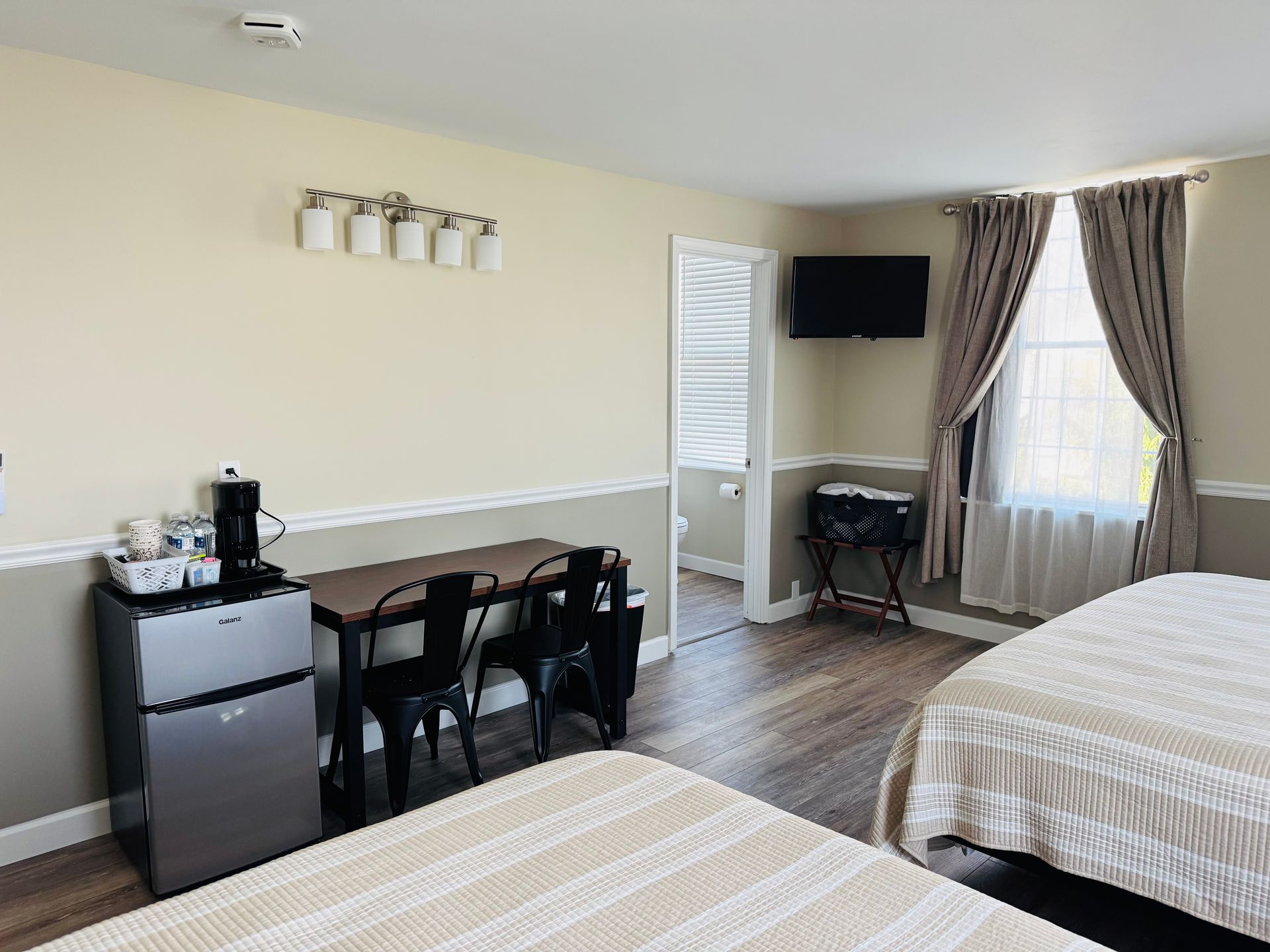 A hotel room with two beds, a small table with two chairs, a mini fridge, a television, and a window with tan curtains.
