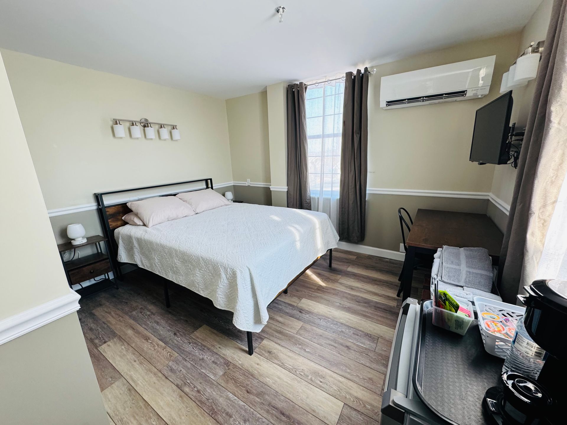 A hotel room featuring a queen bed with white bedding, wood-look flooring, a desk, a television, and a wall-mounted AC unit.