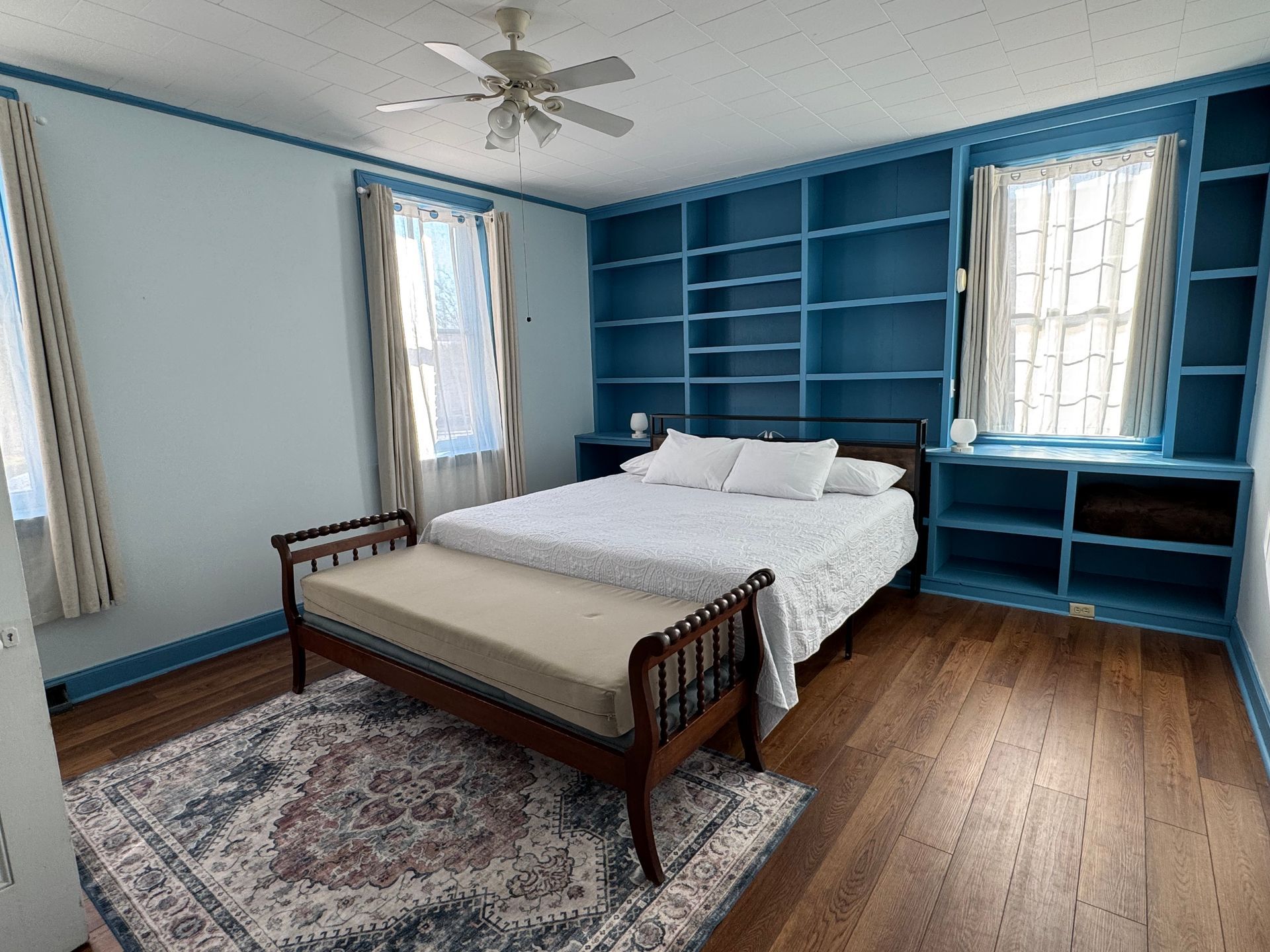 A bedroom with a queen bed, a wood bench at the foot, hardwood floors, a patterned area rug, and built-in blue shelving.