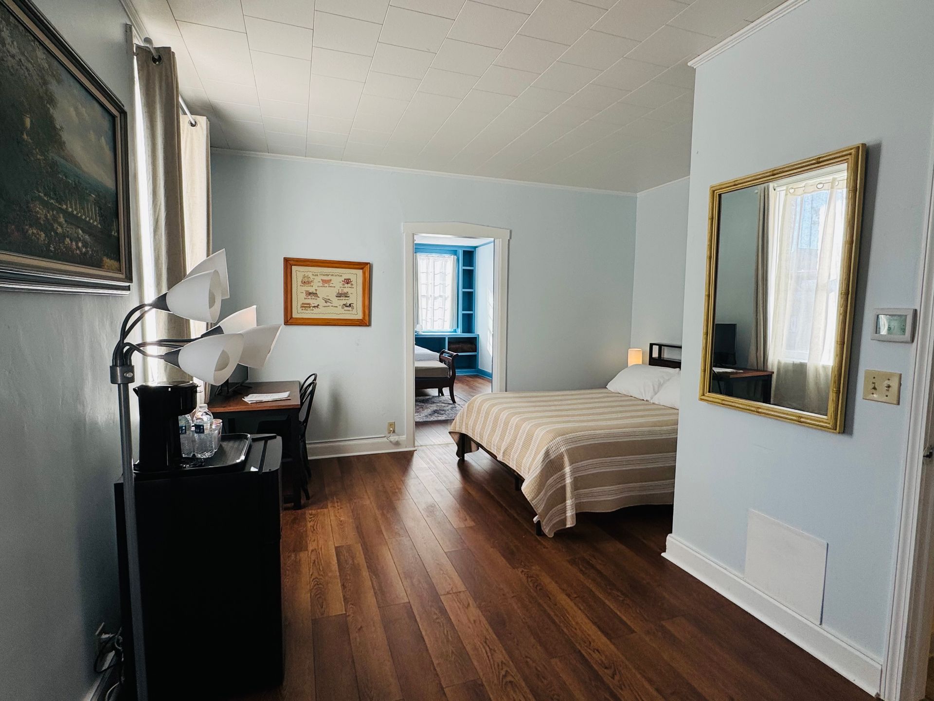 A hotel room with light blue walls, wood flooring, a bed, a desk, a tall lamp, and a large gold-framed wall mirror.