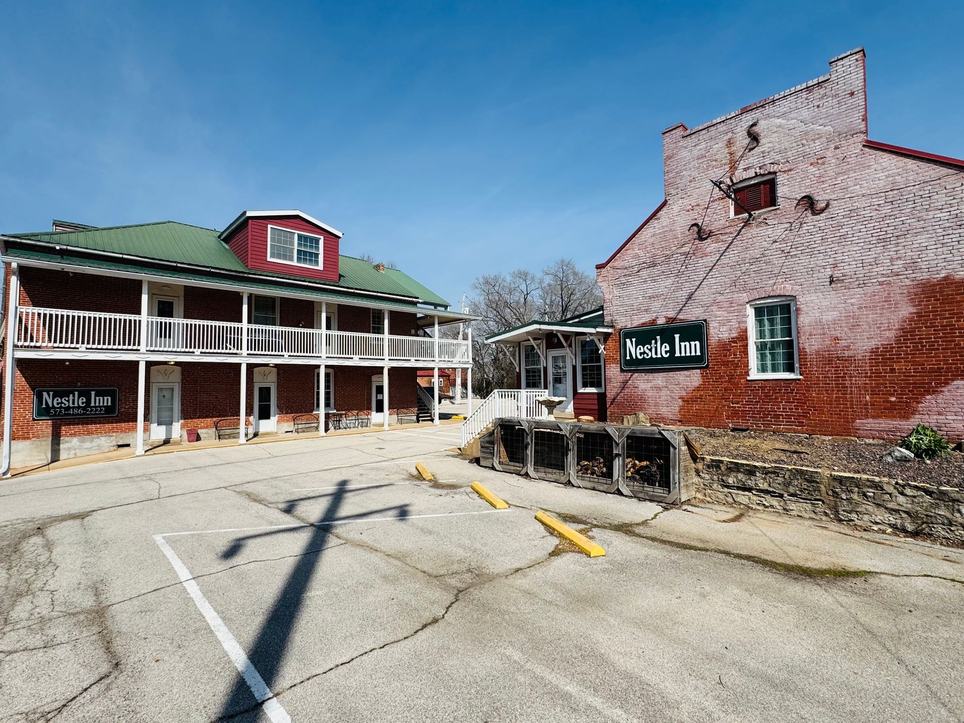 Two historic buildings, a red brick inn and a white-balconied structure, stand in a sunny, paved parking lot.