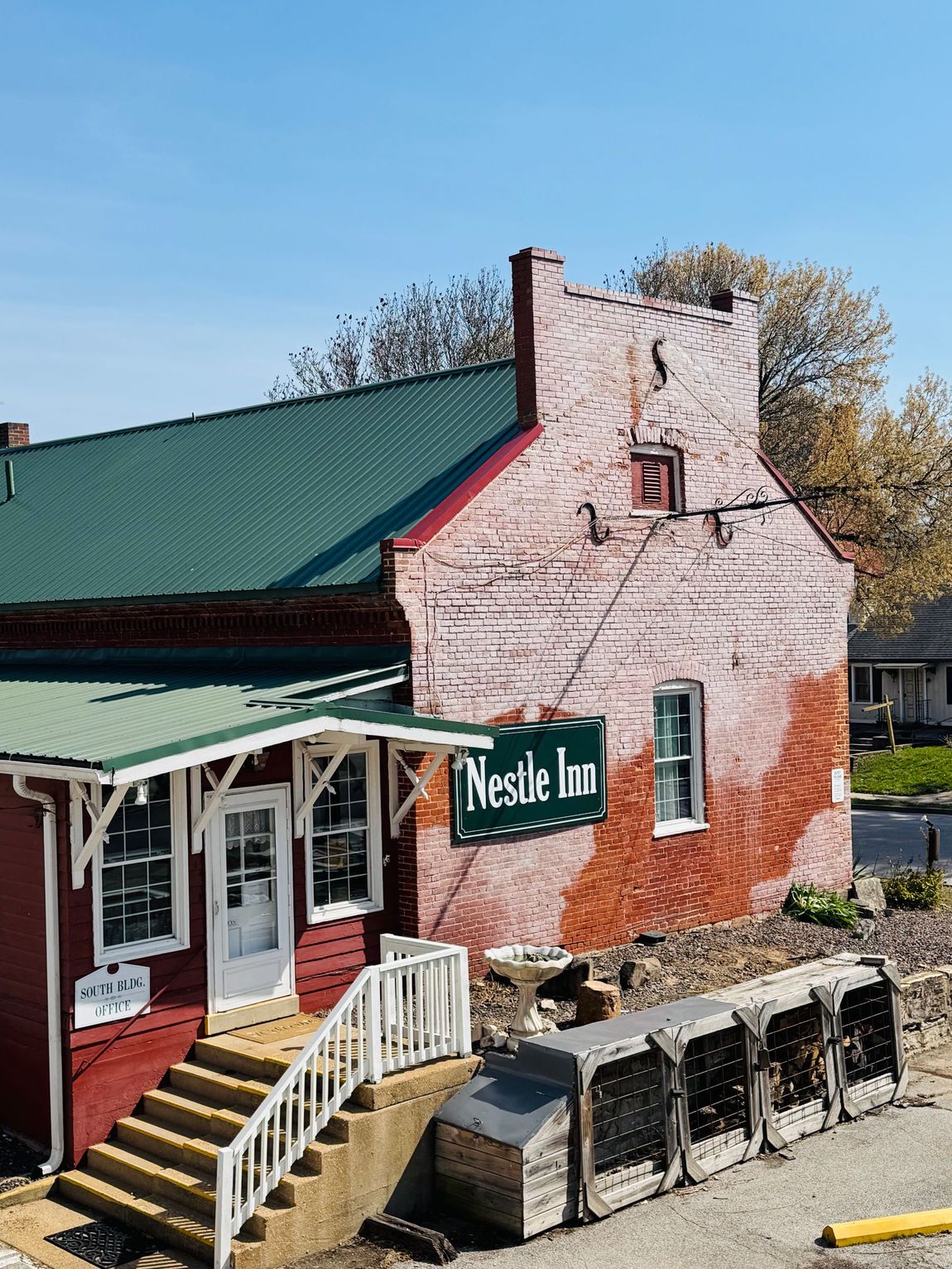 An old red brick building with a green metal roof, a white porch, and a sign that reads 