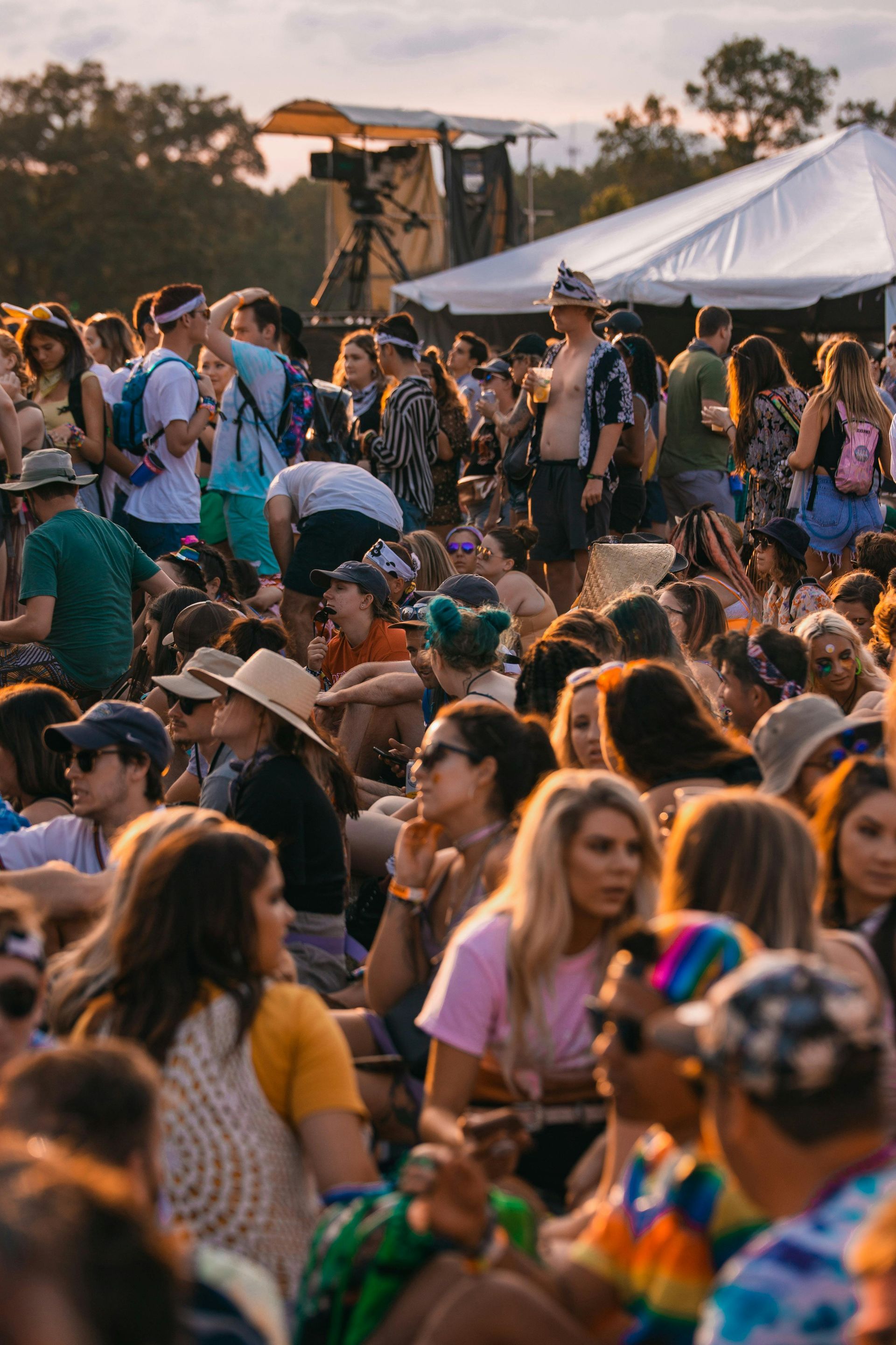 A diverse crowd of people gathered outdoors on a sunny day at a music festival, with tents and equipment in the background.