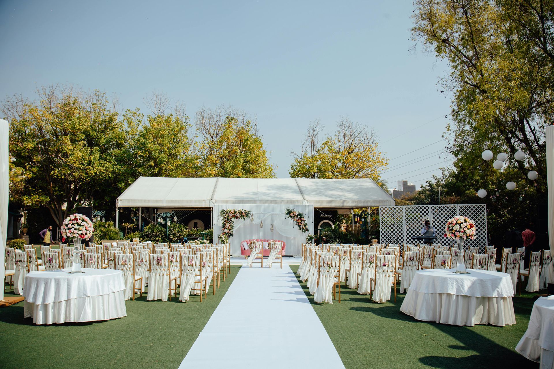 An outdoor wedding venue with a white aisle, rows of chairs, floral arrangements, and round tables on a grassy lawn.