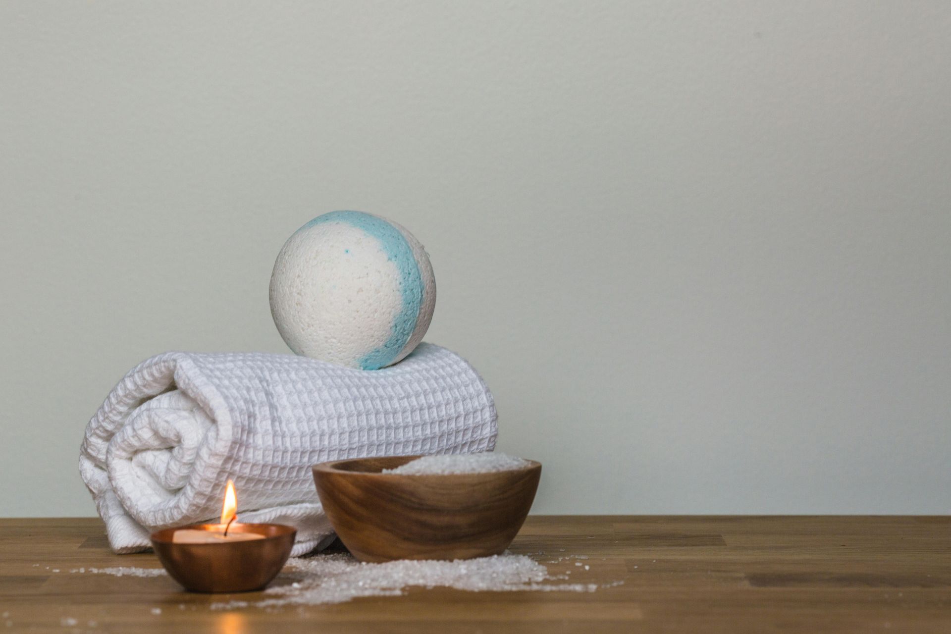 A white rolled towel with a blue-and-white bath bomb, a lit candle, and a bowl of bath salts on a wooden table.