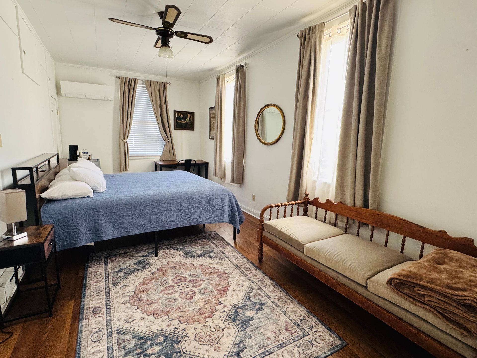 A bedroom with a blue-quilted bed, a wooden bench, a patterned area rug, two windows with curtains, and a ceiling fan.