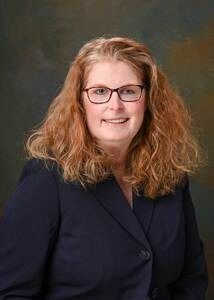 Studio portrait of a person with glasses and curly red hair, wearing a dark blazer and smiling.