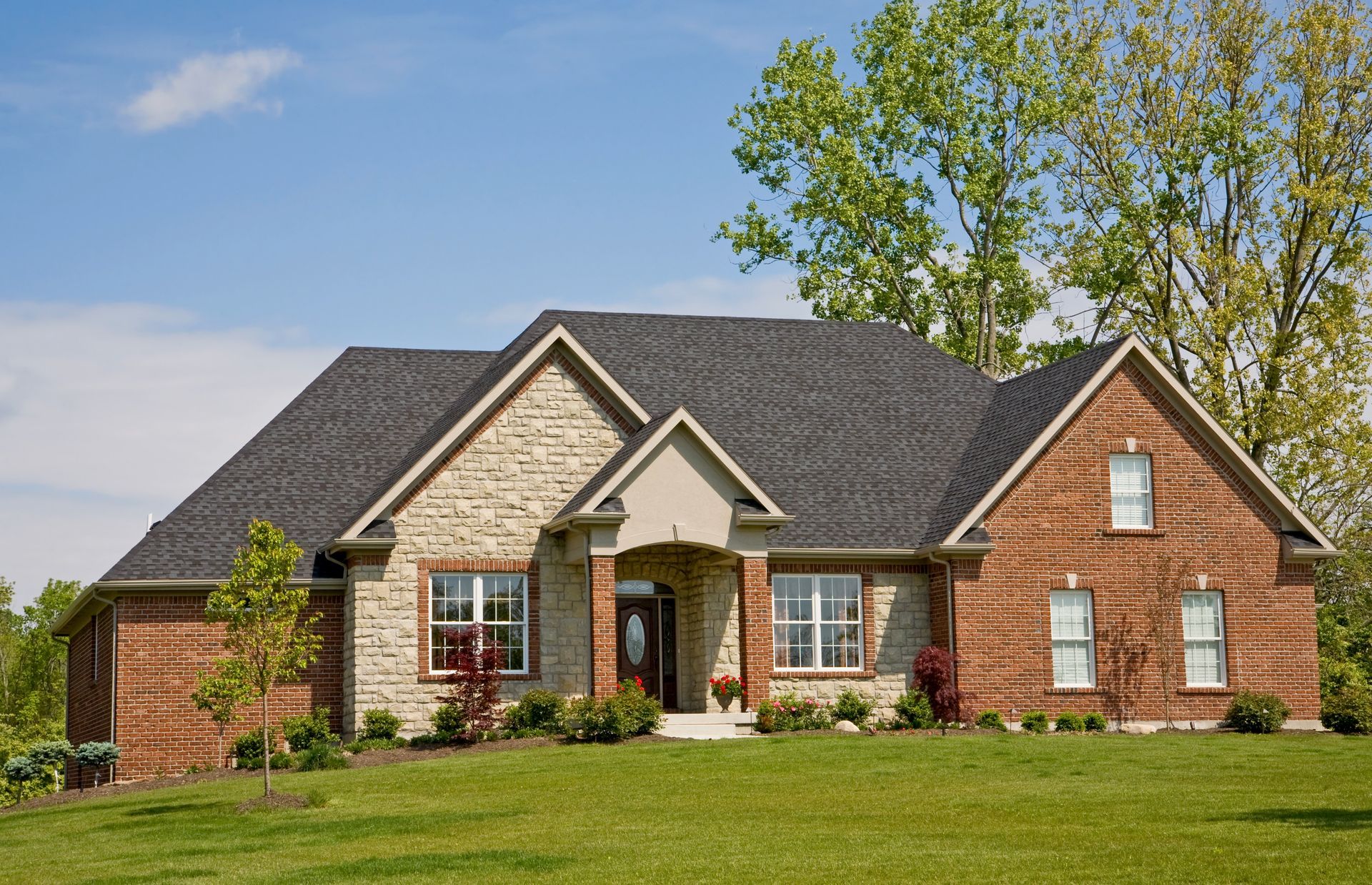 Brick and stone house with dark roof on a grassy lawn under a blue sky.