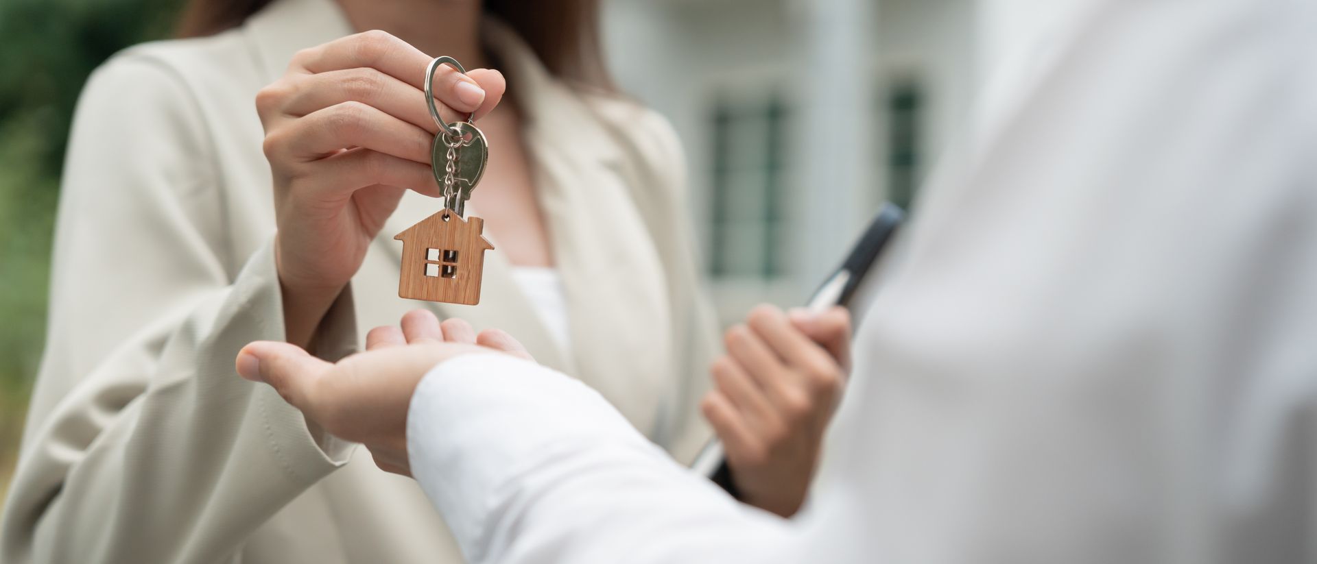 A person hands a key with a wooden house charm to another person, likely a real estate transaction.