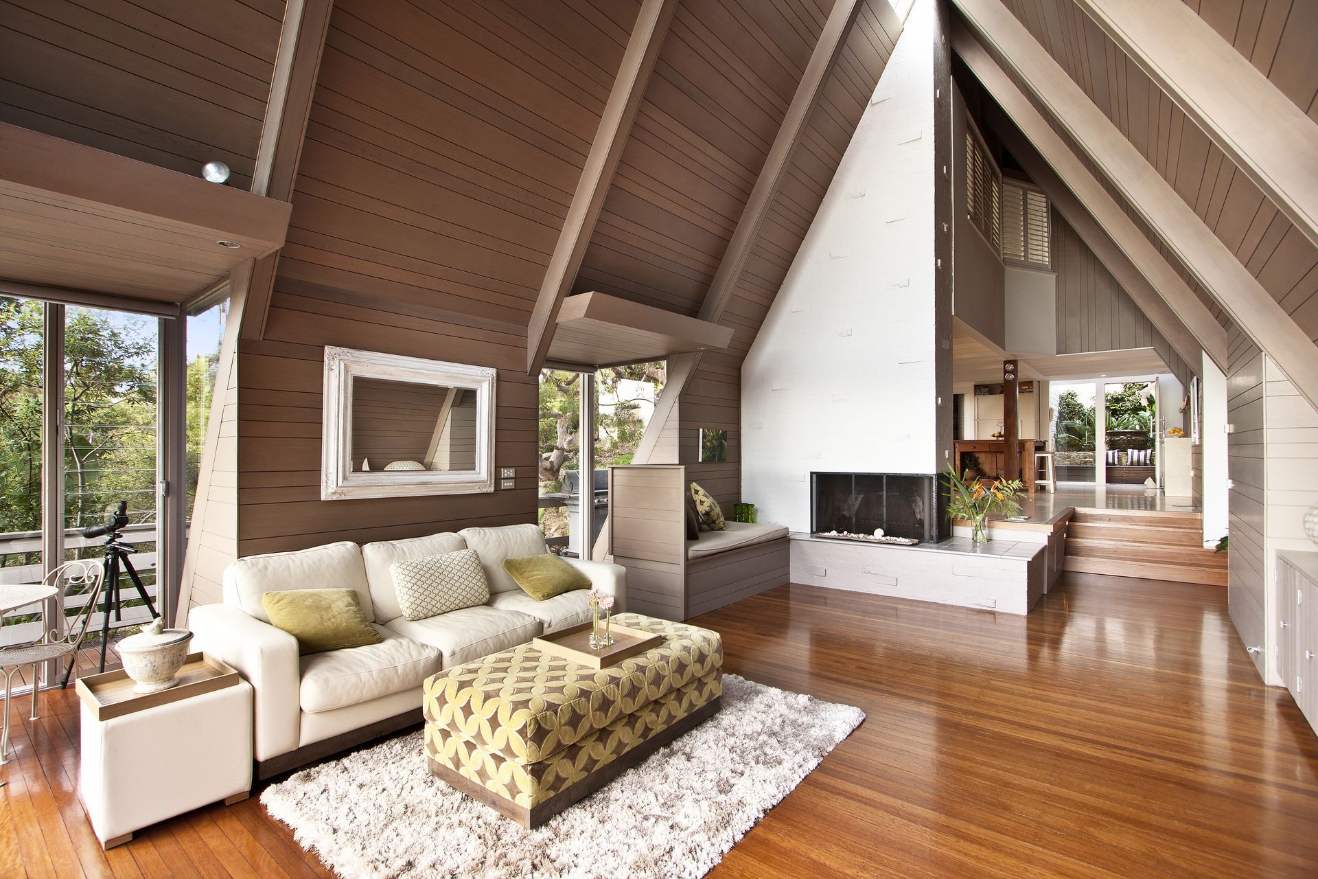 Living room with high, sloped wooden ceiling, white walls, and a fireplace. Furnishings include a sofa, ottoman, and area rug.