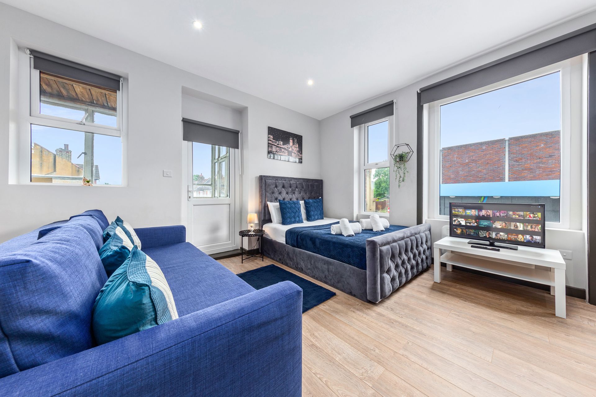 Blue studio apartment with bed, sofa, TV, and large windows.