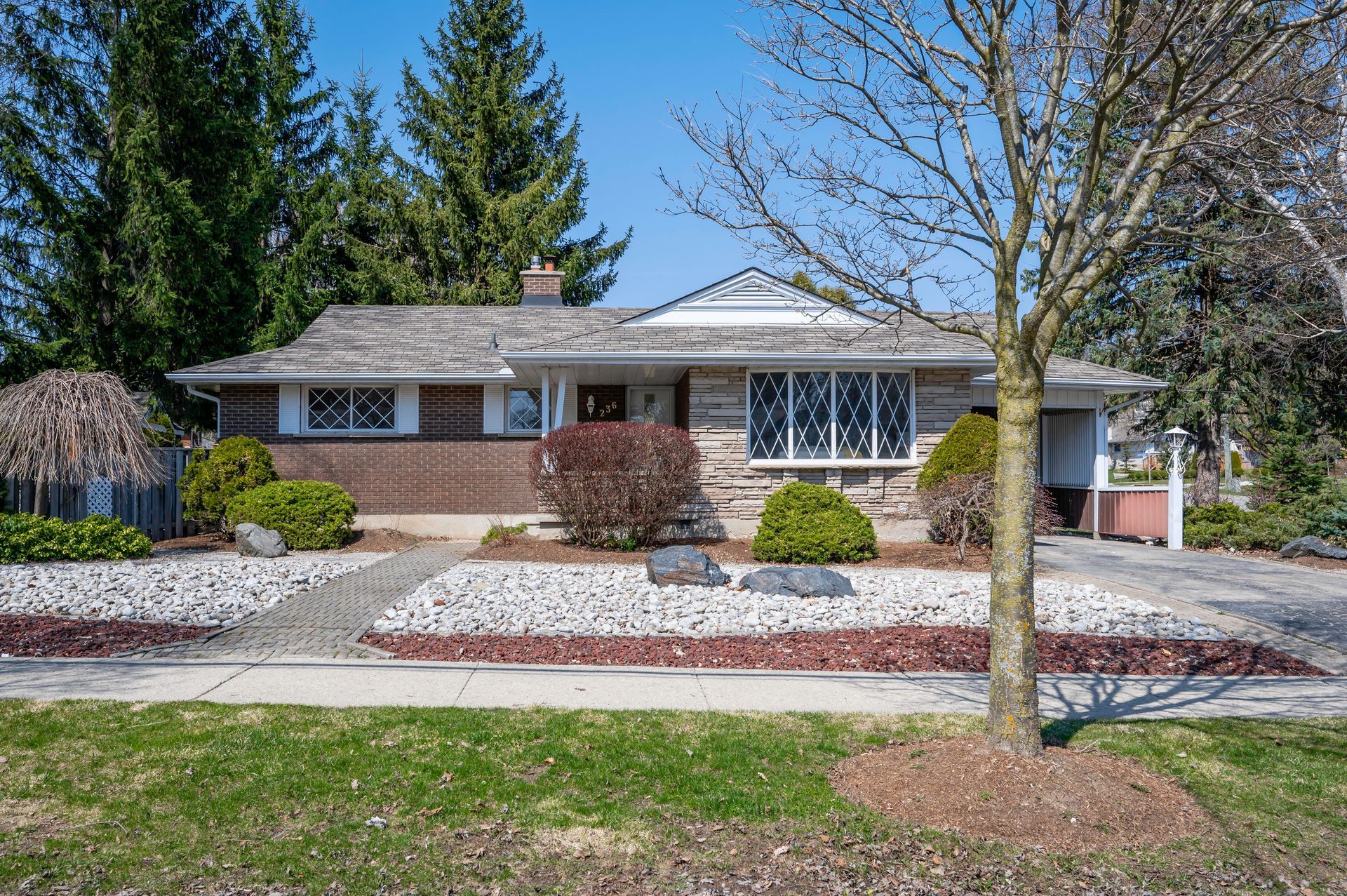 Ranch-style house with stone and brick exterior, landscaped front yard with rock pathway, and a tree on a sunny day.