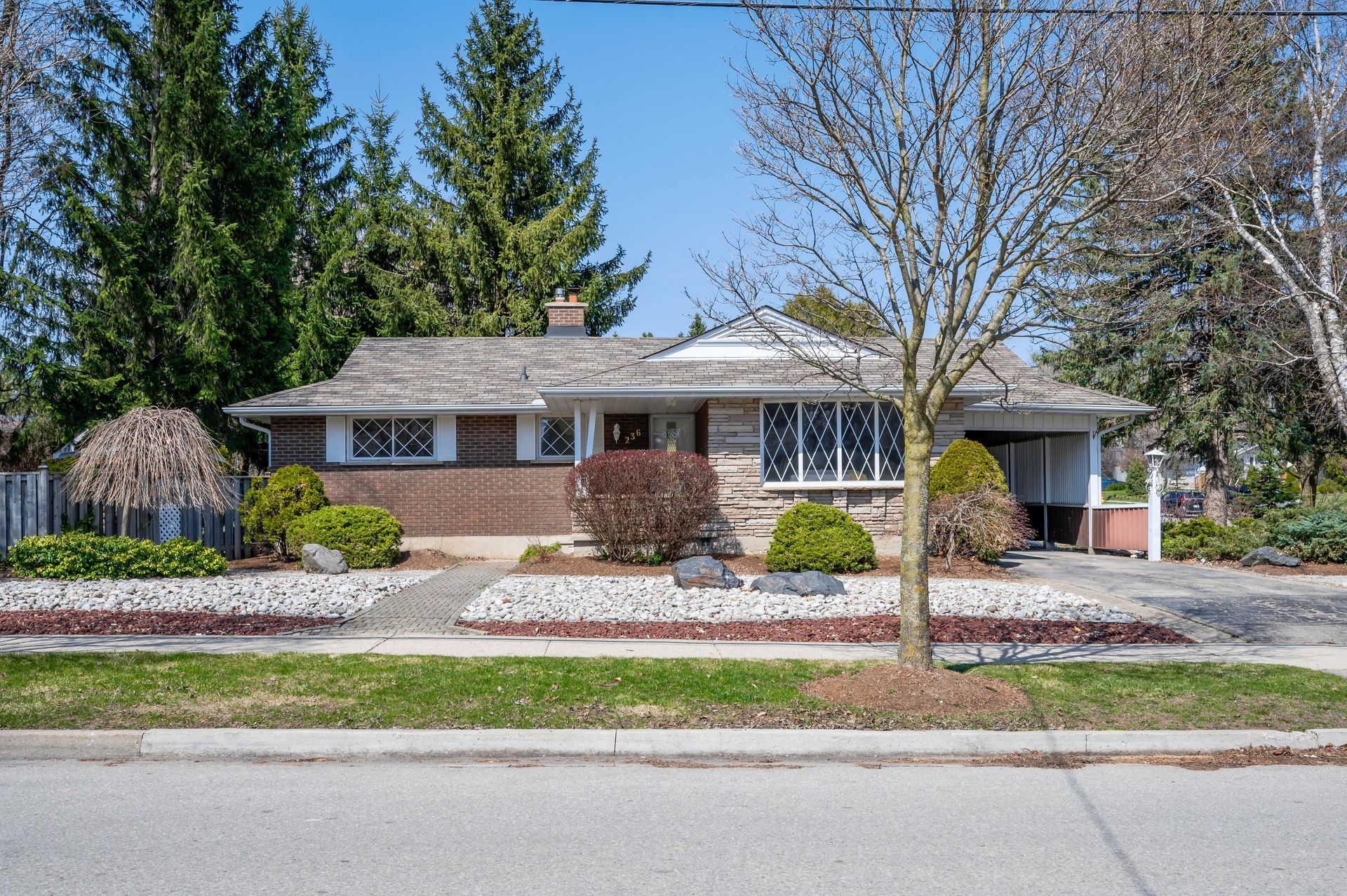 Ranch-style house with brick and stone facade, driveway, small front yard with bushes and a tree. Sunny day.