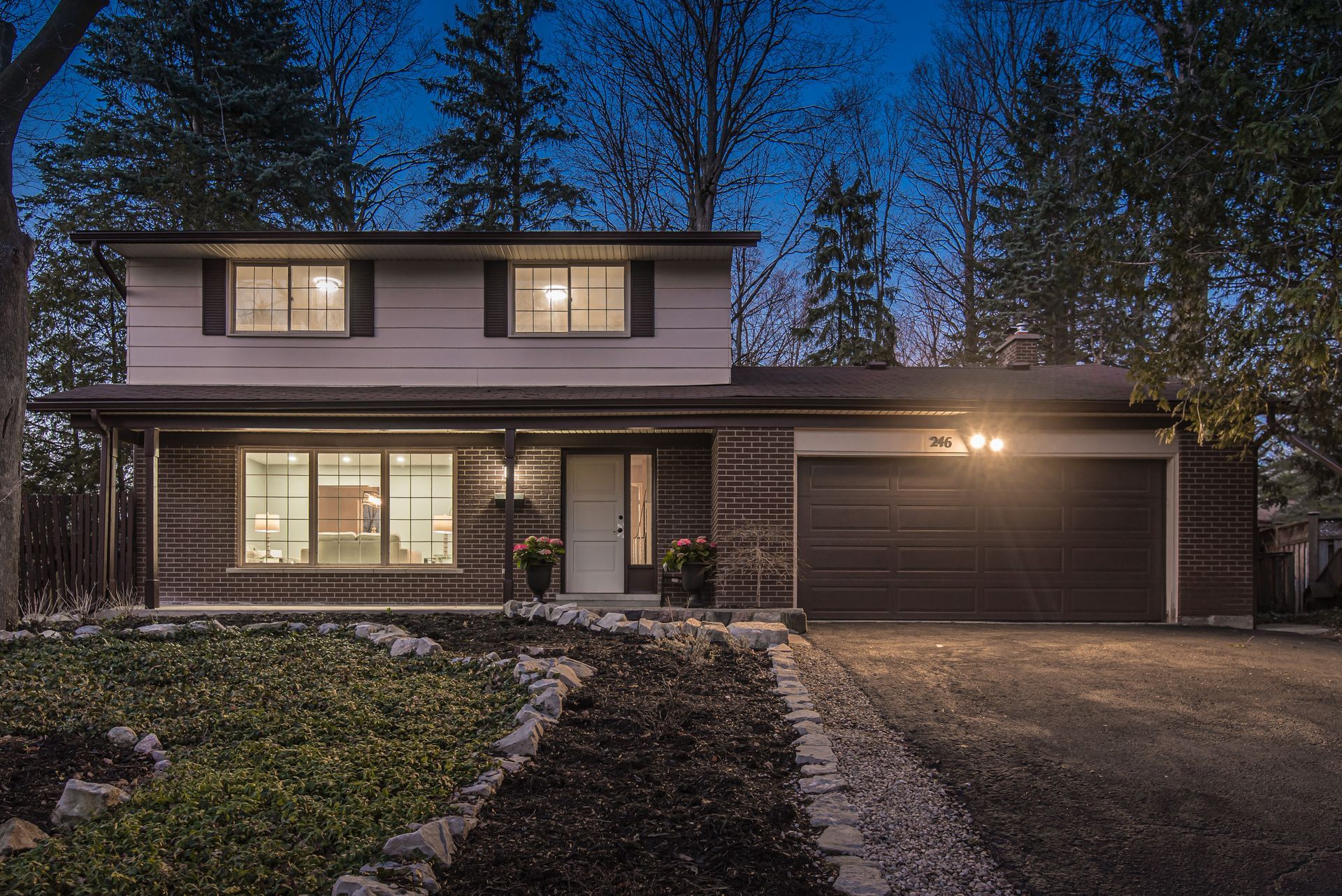 Two-story house with brick facade at dusk; lit windows, garage door, and driveway.
