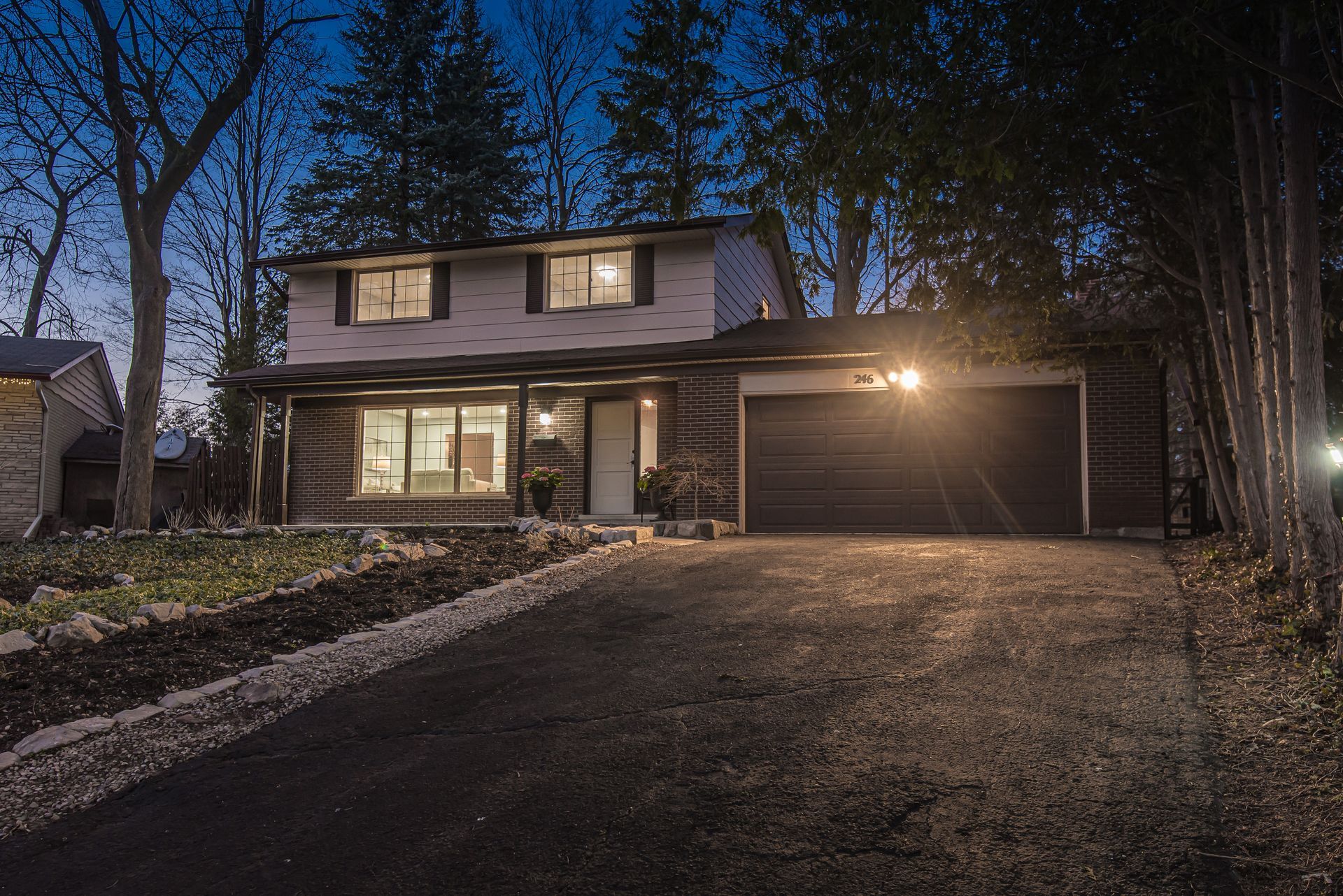 Two-story house with a lit garage and driveway at dusk, surrounded by trees.