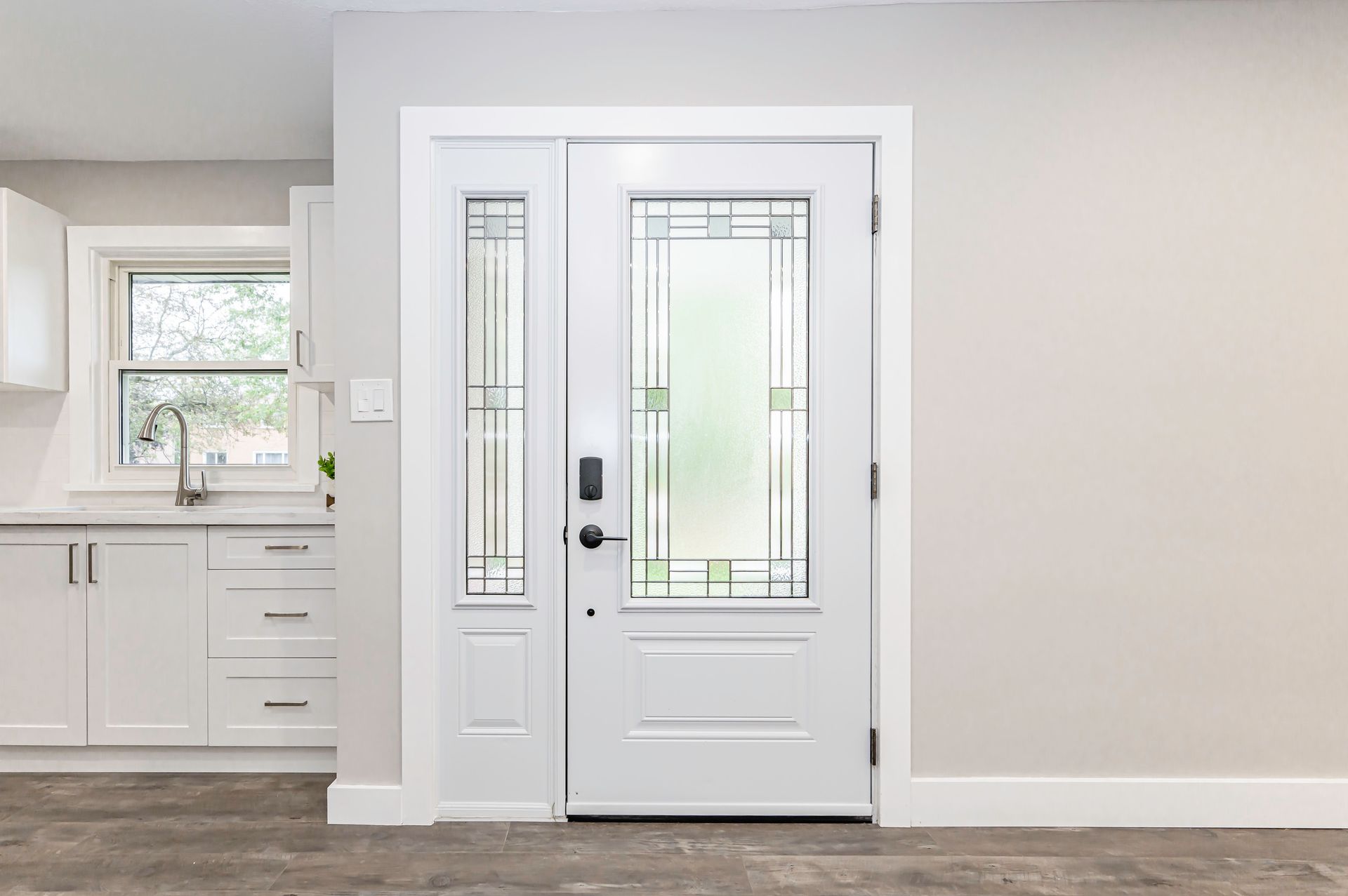 White front door with sidelight and decorative glass, in a neutral-toned room with white cabinets and a window.