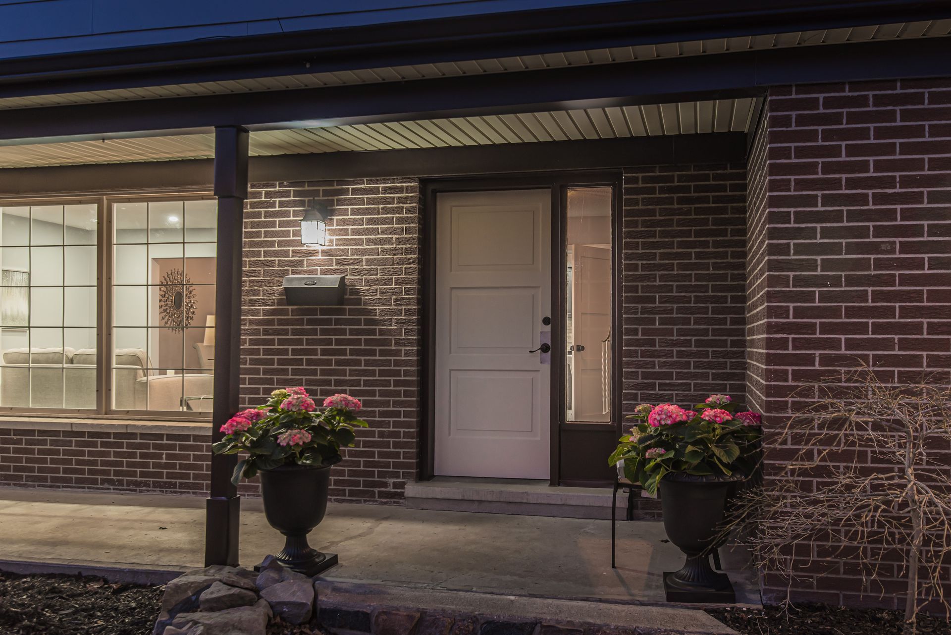 Brick house exterior at dusk, front porch with white door, large windows, and flower pots.